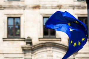 Una bandiera UE collo sfondo un palazzo istituzionale. Immagine di copertina di un articolo di Secondo Welfare sui nessi possibili tra welfare aziendale e Politica di coesione europei, con particolare attenzione sul ruolo delle Regioni.