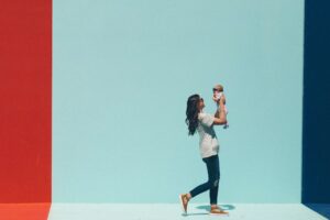 Una mamma tiene in bracco il suo bambino davanti a un muro azzurro, rosso e blu. Immagine di copertina di un articolo di Niccolò Boggian di WhiteLibra sui nessi tra natalità e organizzazione del lavoro