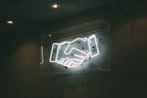 mani che si stringono su una illuminazione al neon. Immagine di copertina di una rticolo di Secondo Welfare in cui si parla del progetto di imapct investing Impact3Coop promosso da Fondazione Social Venture Giordano dell'Amore