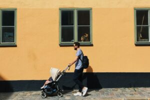 Un uomo spinge un passeggino davanti a una casa gialla. Immagine di copertina di un articolo della serie denatalitalia di Secondo Welfare dedicato alle politiche per la famiglia e la natalità in Svezia.
