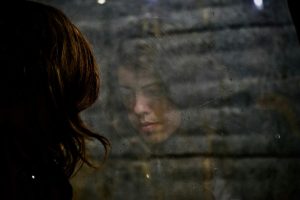 Ragazza alla finestra che guarda il suo riflesso. Immagine di copertina di un articolo di Percorsi di Secondo Welfare sulla salute mentale e l'uso del Budget di Salute