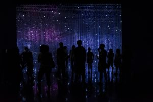 Persone davanti a un muro luminoso fatto di stelle e numeri. Immagine di copertina di un articolo di Percorsi di Secondo Welfare su "Opportunità e rischi di un welfare data-driven nell’era dell’intelligenza artificiale".